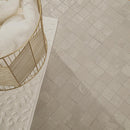 Light beige square mosaic floor tiles shown in a styled interior scene with a white textured bench, a gold wire basket, and white towels.