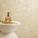 Beige textured square mosaic wall tiles with a white pedestal sink and a rose gold soap dispenser on top, decorated with green eucalyptus leaves.