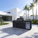 Modern outdoor kitchen with grill and refrigerator in front of a white house with palm trees.