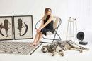 off white foldable metal frame faux leather chair black frame black dressed woman sitting on it dog laying on floor and white side table and 2 picture frames